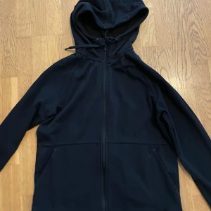 Svart zip hoodie - Svart fleecejacka med hel dragkedja och huva med snörning. Jackan har lång ärm och två sidofickor, samt en något rundad nederkant. Perfekt för chill dagar eller när du vill hålla dig varm. Enkel och stilren design som funkar till det mesta.