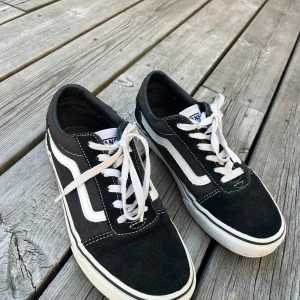 Svarta Vans Old Skool sneakers 37  - Svarta Vans Old Skool sneakers. Svarta m klassisk vit sidorand och vita snören. Material mocka och canvas. Bra skick förutom ett hål vid vristen, Se bild. Innermått 23,5 cm 