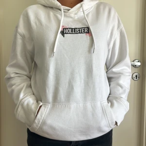 Vit hoodie med blommor från Hollister - Vit hoodie från Hollister i storlek XS med broderade rosa och gröna blommor runt loggan på ryggen. Tröjan har huva med snörning, känguruficka och mjukt material som känns skönt mot huden. Stor i storleken, mer som en storlek M.