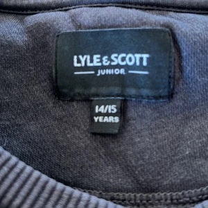 Mörkgrå sweatshirt från Lyle & Scott - Snygg mörkgrå sweatshirt från Lyle & Scott Junior med klassisk rund hals och ribbade muddar. Tröjan har det ikoniska gula Lyle & Scott-logotypen på bröstet och är tillverkad i mjuk bomull. Perfekt för chill dagar och enkel att matcha med jeans eller joggers.