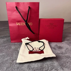 Valentino armband - Snyggt svart armband från Valentino Garavani med justerbar snörning och en silverfärgad V-logga i metall. Armbandet har en tunn, stilren design och en liten oval bricka med Valentino Garavani-gravyr. Perfekt accessoar för att lyfta din outfit.