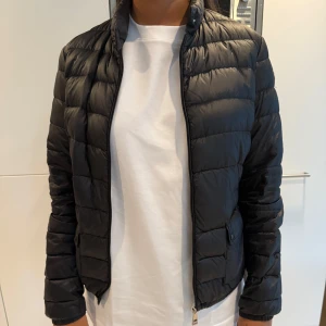 Svart Moncler dunjacka - Snygg svart dunjacka från Moncler med quiltad design och hög krage. Jackan har dragkedja framtill, sidofickor med knapp och Moncler-logga på ärmen. Perfekt för dig som vill ha en stilren och varm jacka med exklusiv känsla.