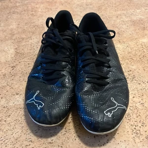 Svarta fotbollsskor från Puma - Säljer ett par svarta fotbollsskor från Puma med vita detaljer och logga på både tå och häl. Skorna har snörning och en rund tå, samt mönstrade prickar över hela ovandelen. Yttermaterialet är syntet och sulan är designad för bra grepp på planen.
