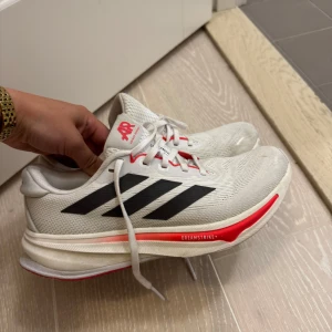Adidas Supernova vita sneakers strl 38⅔ - Säljer ett par Adidas Supernova sneakers i vitt med svarta ränder och röda detaljer. Skorna har mesh-ovandel för bra ventilation, snörning och platt sula med Dreamstrike-dämpning. Perfekta för träning eller löpning, riktigt snygga och sportiga. 