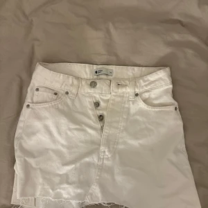 Vit jeanskjol med rå kant från Gina Tricot - Säljer en vit jeanskjol från Gina Tricot i storlek 34. Kjolen har en kort modell med rå, fransig kant nedtill och klassiska fickor fram. Stängs med knappar och har en cool, avslappnad vibe. Perfekt för dig som gillar streetstyle och denim.