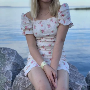 Vit blommig kortklänning med volang - Supersöt vit kortklänning med rosa blommor och puffiga kortärmar. Klänningen har smockad midja och volangkant nedtill som ger en gullig vibe samt ger fina former. Offshoulder-stil och figurnära passform, perfekt för sommarens alla tillfällen. Materialet känns lätt och luftigt🥰. Detta har varit min absoluta favorit klänning så gör ont att sälja den men tyvärr är den för liten