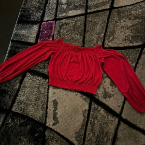 Röd offshoulder topp från Divided. Storlek XS/S.  - Säljer en röd offshoulder topp från Divided i storlek S men kan också passa som XS eftersom den är stretchig. Toppen har långa ärmar, smockad resår vid axlar och midja samt en croppad passform. Materialet är mjukt och lätt, perfekt för att ge en chill och trendig vibe. Snygga volangdetaljer vid kanterna för extra stilpoäng. Jättefin topp och bara använd ett fåtal gånger.😊