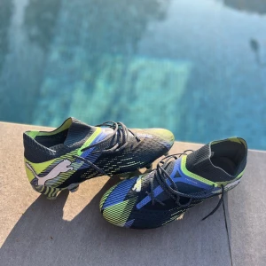 Puma fotbollsskor blå/gul mönster - Säljer ett par grymma Puma fotbollsskor med coolt randigt mönster i blått, svart och neongult. Skorna har snörning, låg profil och en stickad ovandel för skön passform. Sulan har mönstrade dobbar för max grepp på planen. Perfekt för dig som vill sticka ut på matchen!