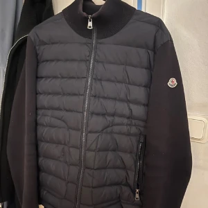 Moncler svart cardigan  - Svart pufferjacka från Moncler med quiltad framsida och släta ärmar. Jackan har ribbad krage, dragkedja framtill och Moncler-logga på ärmen.Jackan har ett litet pyte hål i magen Materialmix med polyester och bomull ger en skön känsla och snygg look. Perfekt för dig som gillar stilrena och moderna jackor.