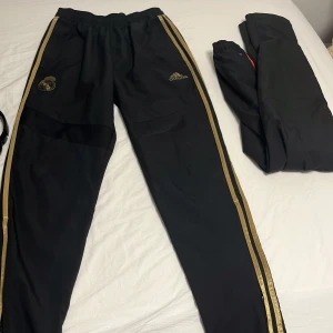 Svarta Real Madrid träningsbyxor Adidas - Snygga svarta träningsbyxor från Adidas med Real Madrid-logga i guld på ena benet och Adidas-logga i guld på andra. Guldiga ränder längs sidorna ger en sportig vibe. Byxorna har elastisk midja och är gjorda i lätt polyester, perfekta för fotboll eller chill.