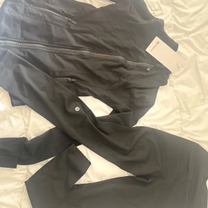 Svart vindjacka från Lululemon - Snygg svart vindjacka från Lululemon med figurnära passform och långa ärmar. Jackan har två praktiska fickor med dragkedja och en stilren design med Lululemon-logga på ärmen. Perfekt för dig som gillar sportig och clean stil.