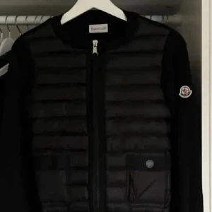 Moncler cardigan  - Svart dunväst från Moncler med quiltad framsida och två fickor med knappstängning. Västen har dragkedja framtill och klassisk Moncler-logga på ärmen. Materialet är dun och polyester, perfekt för kyliga dagar. Snygg och stilren design som funkar till många outfits.