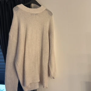 Beige stickad tröja från 157 - Mysig beige stickad tröja från 157 med bred ribbad kant vid hals, ärmslut och nederkant. Tröjan har en loose passform och långa ärmar, perfekt för att hålla sig varm under kyliga dagar. Enkel och stilren design som är lätt att matcha med jeans eller leggings.