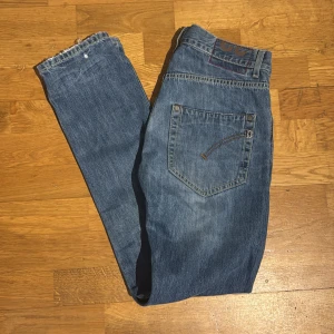 Dondup blå jeans, storlek 32 - Säljer ett par dondup jeans i blå tvätt med raka ben och knappgylf. Jeansen har fem fickor, snygga slitningar och detaljer på bakfickorna. Tillverkade i 100% bomull för en autentisk jeanskänsla. Perfekt för dig som gillar tidlös streetstil.