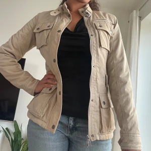 Beige parkajacka med fickor och dragkedja - Snygg beige parkajacka med hög krage, spänne och både knappar och dragkedja framtill. Jackan har långärmade ärmar och en figurnära passform.