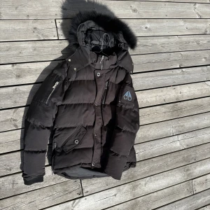Svart dunjacka från Moose Knuckles - Säljer en svart dunjacka från Moose Knuckles med fluffig päls på huvan, coola dragkedjor på ärmarna och flera fickor. Jackan har quiltad design och logga på ärmen. Perfekt för kalla dagar när du vill hålla stilen. Frågor eller funderingar så hör va er!