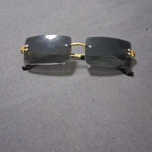 Svarta rektangulära solglasögon med gulddetaljer - Snygga rektangulära solglasögon med svarta linser och tunna guldfärgade metallbågar. Glasögonen har minimalistisk design utan ram runt linserna och svarta skalmar. Perfekt för dig som gillar en stilren och modern look.