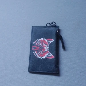 Svart plånbok med röd rävtryck - Cool svart plånbok med ett unikt rävtryck i rött och vitt på framsidan. Plånboken har flera kortfack på baksidan och en praktisk dragkedja för mynt eller småsaker. Materialet är skinn och designen är stilren med en edgy touch.