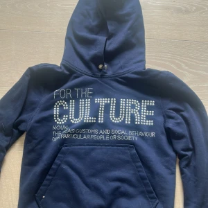 Mörkblå hoodie med pärltext - Säljer en mörkblå hoodie med texten 'FOR THE CULTURE' i pärlor på bröstet. Tröjan har huva med knappdetalj, magficka och långa ärmar. Materialet känns som mjuk bomull och passformen är normal. Perfekt för dig som gillar statement-plagg och coola detaljer.