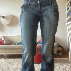 Blå bootcut jeans från Mouni Jeanswear - Snygga blå jeans med bootcut från Mouni Jeanswear. De har låg midja, slitna detaljer och unika bakfickor med broderad kant. Jeansen är tillverkade i klassiskt denim och har en cool tvättad look med ljusare partier framtill och baktill. Stretchigt tyg .