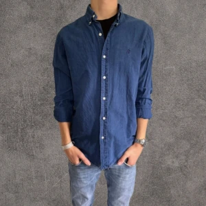 Blå oxfordskjorta från Ralph Lauren slimfit - Slim fit oxfordskjorta från Ralph Lauren i en snygg mörkblå färg. Skjortan har klassisk button down-krage, vita knappar och broderad logga på bröstet. Tillverkad i mjuk bomull och passar perfekt till jeans eller chinos.