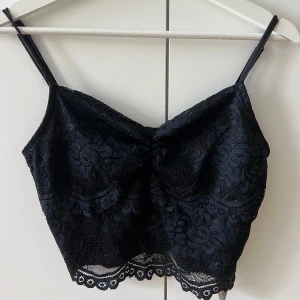 Svart spetsig croppad linnetopp - Supersnygg svart croppad linnetopp med tunna axelband och blommig spets över hela. Toppen har en lätt v-ringad form och är fodrad på insidan för extra komfort. Perfekt att styla med jeans eller kjol för en cool look.