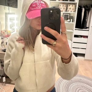 Vit croppad hoodie med dragkedja - Säljer en vit croppad hoodie med huva och dragkedja framtill. Tröjan har långa ärmar, ribbade muddar och en stor ficka på magen. Mjuk stickad känsla som är perfekt för chill dagar. Enkel och clean look som funkar till jeans eller mjukis.