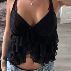 Svart volang blus med spetsdetaljer - Säljer en svart blus med tunna axelband och snygga volanger i lager på framsidan. Toppen har spetsdetaljer och en knapp framtill. Den är aldrig använd💕