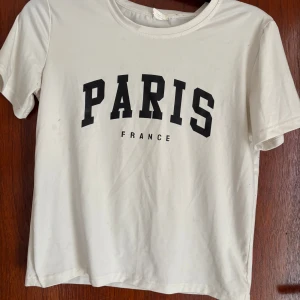 Vit t-shirt med PARIS-tryck, S - Vit t-shirt i storlek S med svart text 'PARIS FRANCE' på bröstet. Klassisk rund hals och korta ärmar. Tillverkad i 100% polyester för en lätt och mjuk känsla. Perfekt för dig som gillar enkel och stilren streetwear med en touch av Paris.