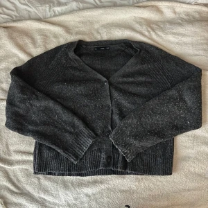 Mörkgrå stickad kofta från MNG - Mysig mörkgrå stickad kofta från MNG med v-ringning och knappar framtill. Koftan har långa ärmar och bred ribbad mudd vid ärmslut och nederkant. Bra skick men noppig 