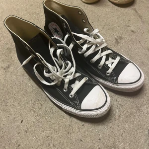 Svarta Converse Chuck Taylor High - Klassiska svarta Converse Chuck Taylor All Star high tops med vit tå och vita snören. Skorna har rund tå, platt sula och tydliga vita sömmar. Märket syns på sidan och insidan är beige. Ovandel och foder i textil, yttersula i gummi.