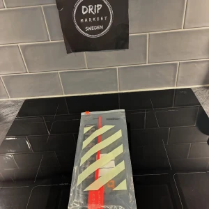 svart off white nyckelring  - säljer en exklusiv svart off white nyckelring med en tydlig logotyp i grått och rött. nyckelringen har en cool zip-tie i röd plast och metallspänne. kommer med i originalförpackning med off white klassiska randiga design på påsen. perfekt för dig som gillar streetwear och vill sticka ut.