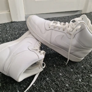 Vita Nike high sneakers - Fräscha helvita Nike sneakers med högt skaft och klassisk swoosh på sidan. Skorna har snörning och är tillverkade i syntetmaterial och skinn. Perfekta för dig som gillar en clean och stilren look.