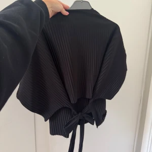 Svart ribbad topp med knyt bak - Säljer en svart ribbad topp med vida ärmar och snygg knytning i ryggen. Toppen har en boxig, loose passform och är tillverkad i ett mjukt, stretchigt material. Perfekt för dig som gillar stilrena plagg med en twist.