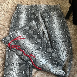 Grå bootcut byxor med ormmönster - Säljer ett par grå bootcut byxor med snyggt ormmönster med en röd logo ”Kani” längs benet. Byxorna har hög midja och är tillverkade i ett mjukt material som sitter skönt. 