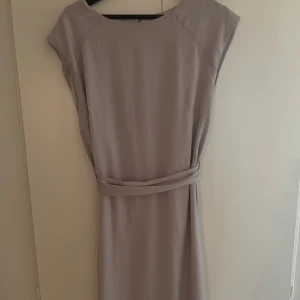  långklänning  - Långklänning med rund halsringning och avtagbart bälte i midjan. Klänningen har ärmlös design och knäppning bak i nacken. 