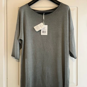 Grågrön tunika/klänning från Cotton Candy - Enkel och stilren grågrön Tunika/tröja från Cotton Candy. Modellen är rak med rund halsringning och halvkort ärm. Färgen kallas Dusty Olive och materialet är mjukt och bekvämt, perfekt för en chill look.