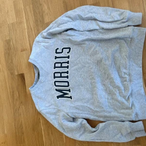 Grå sweatshirt från Morris, storlek L - Snygg grå sweatshirt från Morris med stor svartvit logga över bröstet. Klassisk rund halsringning och ribbade muddar vid ärmslut  nederkant. 
