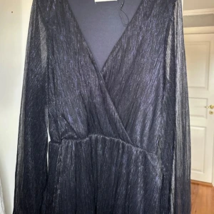 Svart glittrig omlottklänning - Svart långärmad klänning med glittrigt tyg och omlott-skärning framtill. Klänningen har v-ringning och är tillverkad i ett lätt, transparent material som ger en snygg layering-effekt. Perfekt för dig som vill sticka ut med en elegant och trendig look.