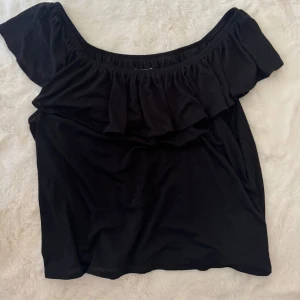 Svart offshoulder topp från H&M - Svart offshoulder topp från H&M med volang längs urringningen. Toppens axlar är bara och den har en avslappnad passform. Perfekt för dig som gillar en enkel men ändå trendig look. Gjord i mjuk bomull som känns skön mot huden.