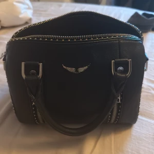 Svart handväska med silverdetaljer - Cool svart handväska från Zadig & Voltaire i skinn med silverfärgade metalldetaljer och dragkedjor. Väskan har två rejäla handtag och en ikonisk vinglogga framtill. Perfekt för dig som gillar edgy och stilren design.
