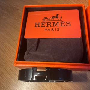 Svart HERM Clic H armband - Säljer ett stilrent svart HERM Clic H armband i metall med emaljdetaljer. Armbandet har den ikoniska H-loggan framtill och kommer i originalask med tillhörande påse och certifikat. Perfekt accessoar för dig som gillar lyxiga detaljer.