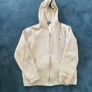 Grå hoodie från Polo Ralph Lauren - Klassisk grå hoodie från Polo Ralph Lauren med dragkedja framtill och broderad logga på bröstet. Tröjan har huva, kängurufickor och är tillverkad i mjuk bomullsblandning. Perfekt för chill dagar eller när du vill ha en clean och enkel look.