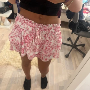 Vit kortkjol med röd blommigt mönster - Supersöt vit kortkjol med rött blommigt mönster och volanger. Kjolen har bred smock i midjan med snörning och är gjord i ett lätt och luftigt material, perfekt för varma dagar. Passformen är normal och kjolen har en feminin och lekfull vibe.