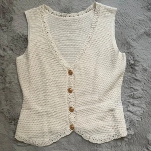 Vit stickad väst med knappar - Superfin vit stickad väst med snygga virkade detaljer längs kanterna och fyra guldiga knappar framtill. Västen har v-ringning och en lätt figursydd passform. 