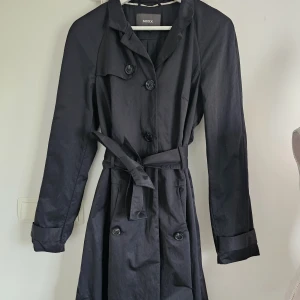 Svart trenchcoat från Mexx med bälte - Stilren svart trenchcoat från Mexx i storlek UK14. Jackan har klassisk krage, knappar framtill och ett matchande bälte i midjan. Långa ärmar med snygga manschetter och slät finish. Perfekt för dig som gillar en tidlös look med lite edge.