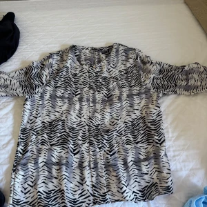 Zebramönstrad blus från Albina - Säljer en luftig zebramönstrad blus från Albina i svart, grått och vitt. Blusen har rund halsringning och långa ärmar, perfekt för dig som gillar djurmönster och vill sticka ut lite extra. Tillverkad i ett mjukt och lätt material.