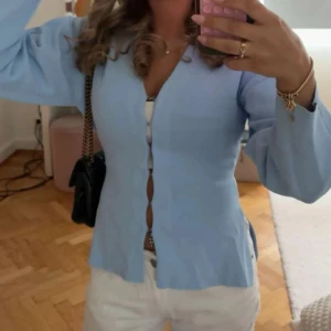SÖKER!! - Supersöt ljusblå blus med v-ringning och knappar framtill. Blusen har långa ärmar med lätt utsvängd form och sitter figurnära över kroppen. Perfekt att styla med jeans eller kjol för en fräsch look.