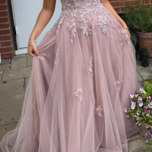Rosa balklänning  - Säljer en rosa långklänning med vacker spets och broderade blommor på livet och kjolen. Klänningen har öppen rygg med snörning och tunna axelband som korsas. Kjolen är i flera lager tunn tyll som ger ett svävande intryck, perfekt för bal.