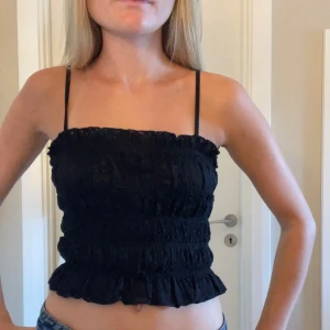 Svart smockad linnetopp  - Supersöt svart linnetopp med smala axelband och smockad struktur. Toppen har volangkant nertill och runt bysten. Oanvänd med prislappen kvar, nypris 199 kr